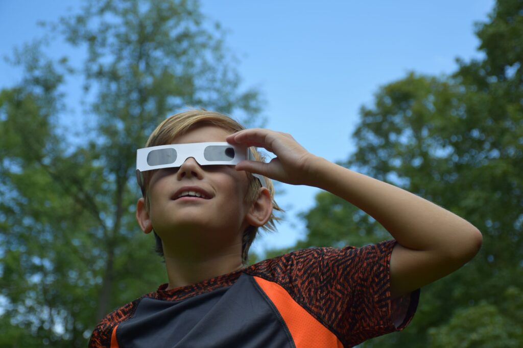 A young boy stands outside and looks at the solar eclipse using special glasses.
